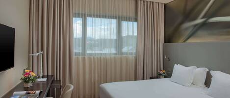 A hotel room with a large bed, a desk with a chair, a flat-screen TV, and a window with curtains.