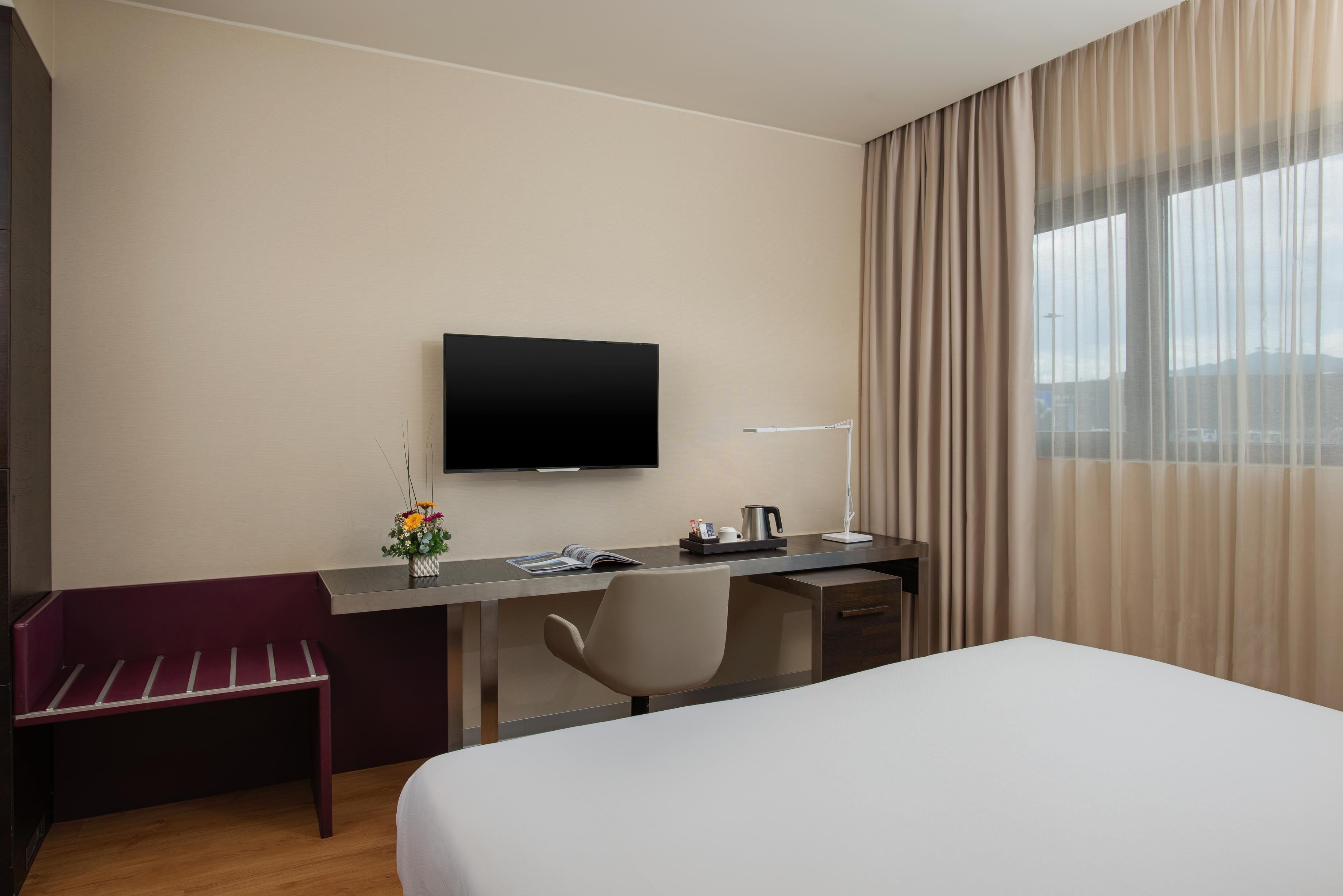 Une chambre d’hôtel comprenant un lit, un bureau avec une chaise, un téléviseur à écran plat, une fenêtre avec des rideaux et un petit banc.