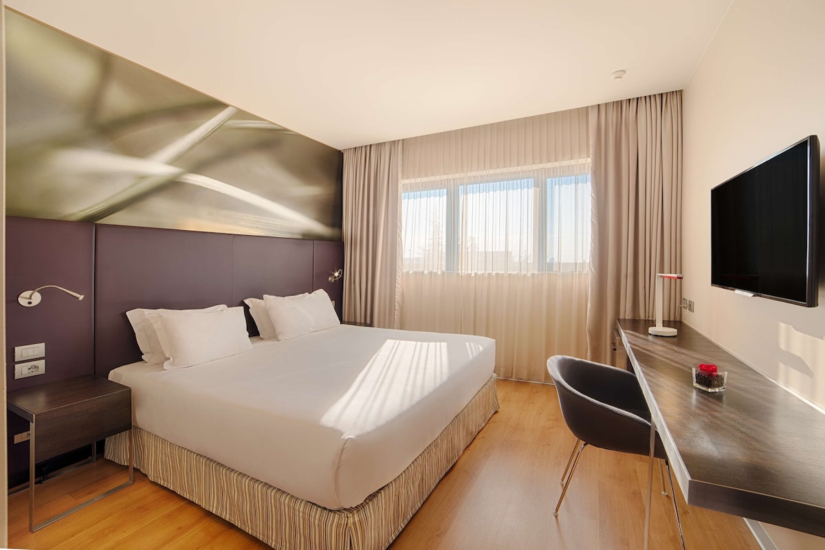 A modern hotel room with a large bed, a desk, a chair, and a TV.
