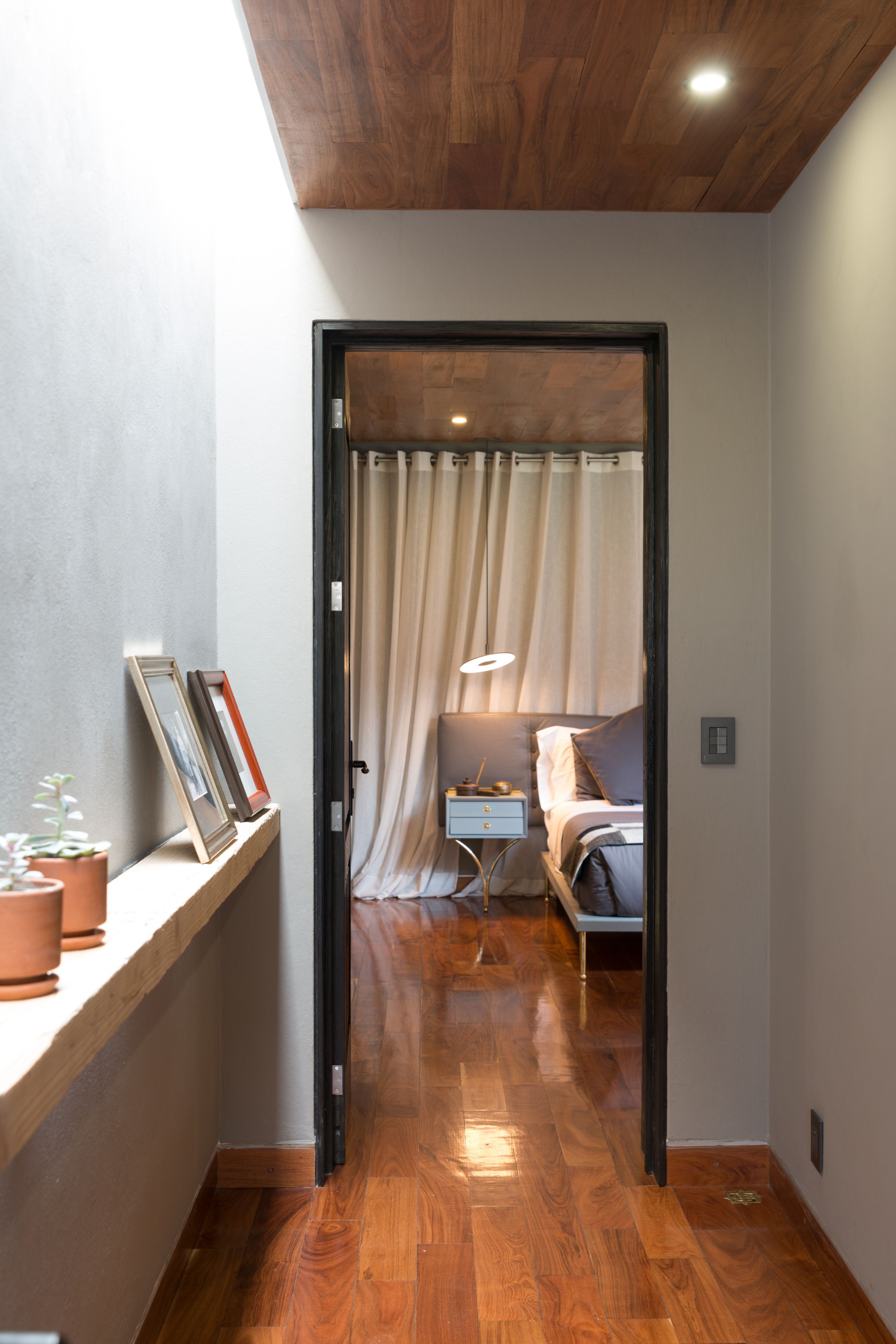 A hallway leading to a bedroom with a bed, bedside table, and a lamp. There is a shelf with framed pictures and potted plants on the left.