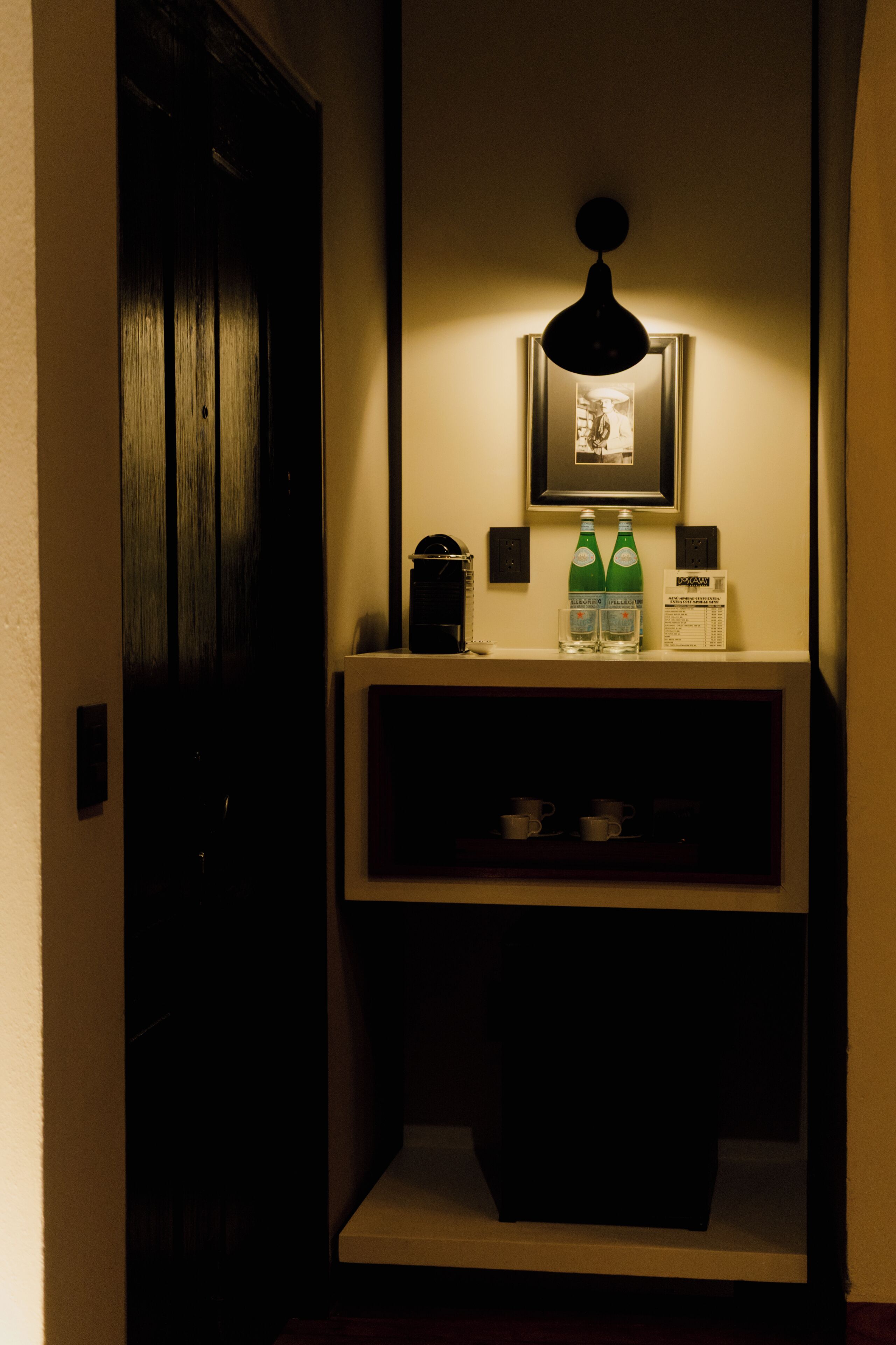 Una habitación de hotel con una puerta de madera oscura, un cuadro en la pared, un estante pequeño con dos botellas y vasos, y una lámpara de pared.