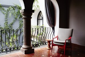 Ein Balkon mit einem roten Stuhl, einer Steinsäule und Blick auf Grün hinter einem Metallgeländer.