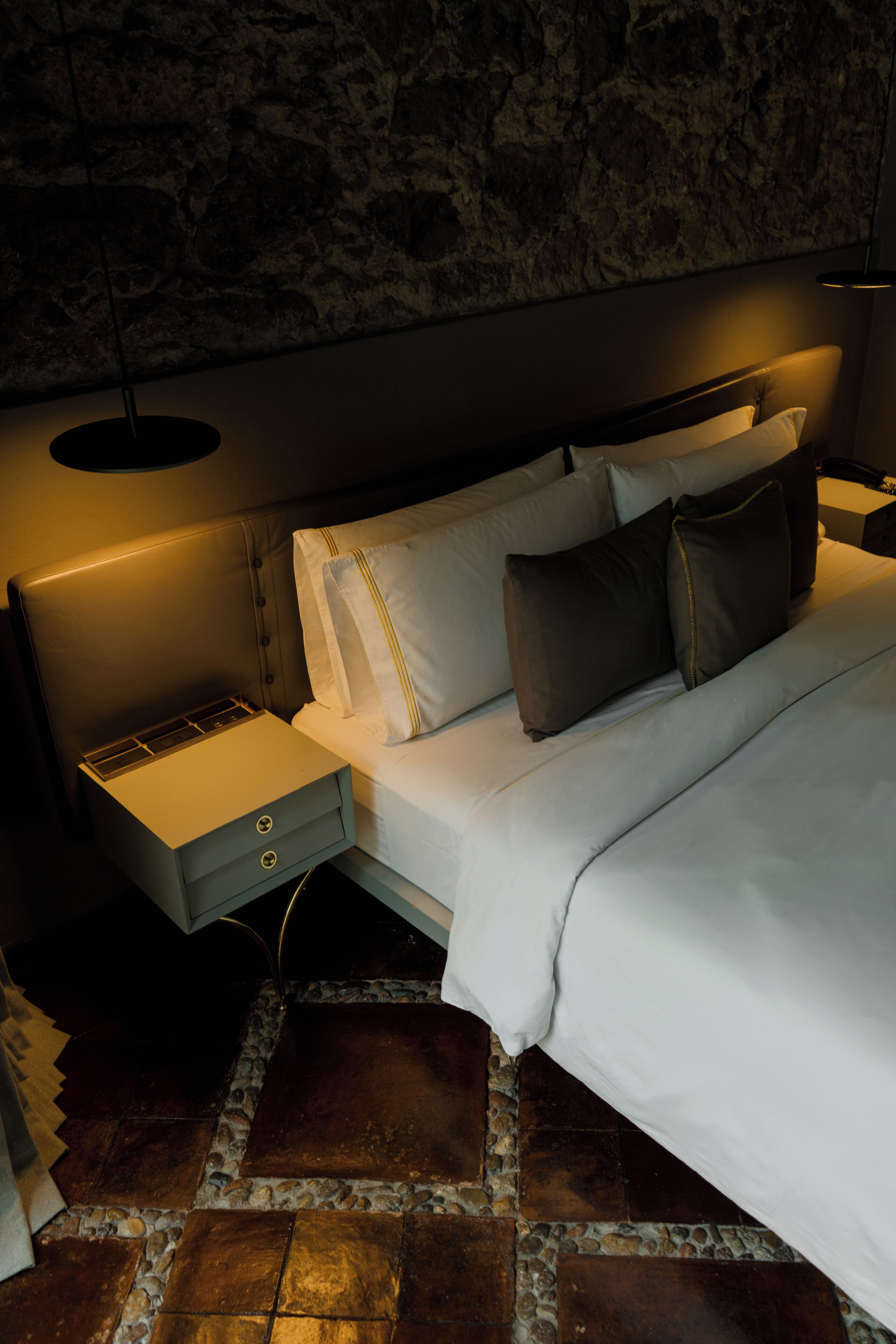 A modern bedroom with a stone wall, a bed with white bedding, a nightstand, and a lamp.