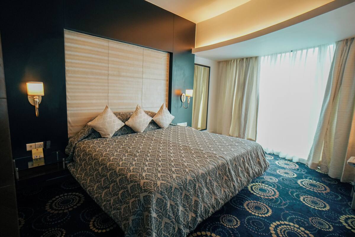 Habitación de hotel con una cama grande, una mesita de noche, una lámpara, una ventana con cortinas y una luz empotrada en la pared.
