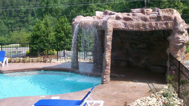 A swimming pool with a waterfall feature, surrounded by a stone wall and a fence.