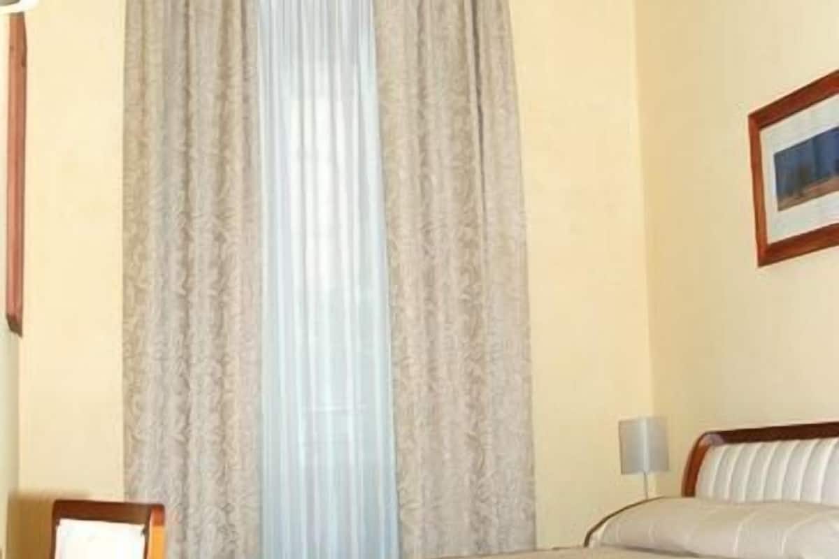 Habitación de hotel con cama, ventana con cortinas, lámpara y un cuadro en la pared.