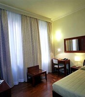 A hotel room with a bed, a desk with a chair, a lamp, and a large window with curtains.
