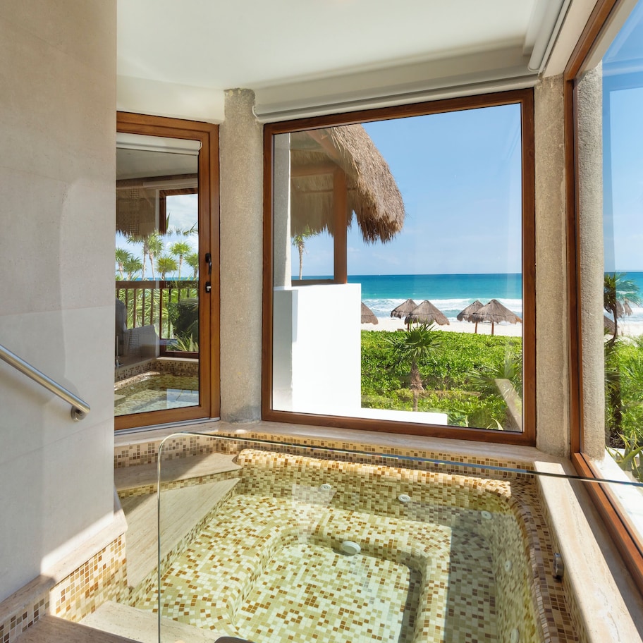 A bathroom with a glass-enclosed jacuzzi, a view of the beach, and a thatched-roof structure.
