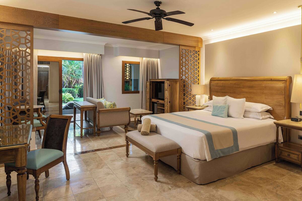A hotel room with a large bed, a desk, a chair, and a bench. There is a ceiling fan, a TV, and a lamp on the bedside table.