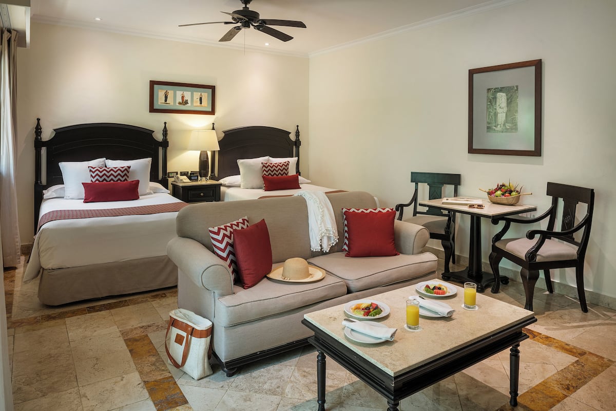 A hotel room with two beds, a sofa, a dining table with breakfast, and a chair.