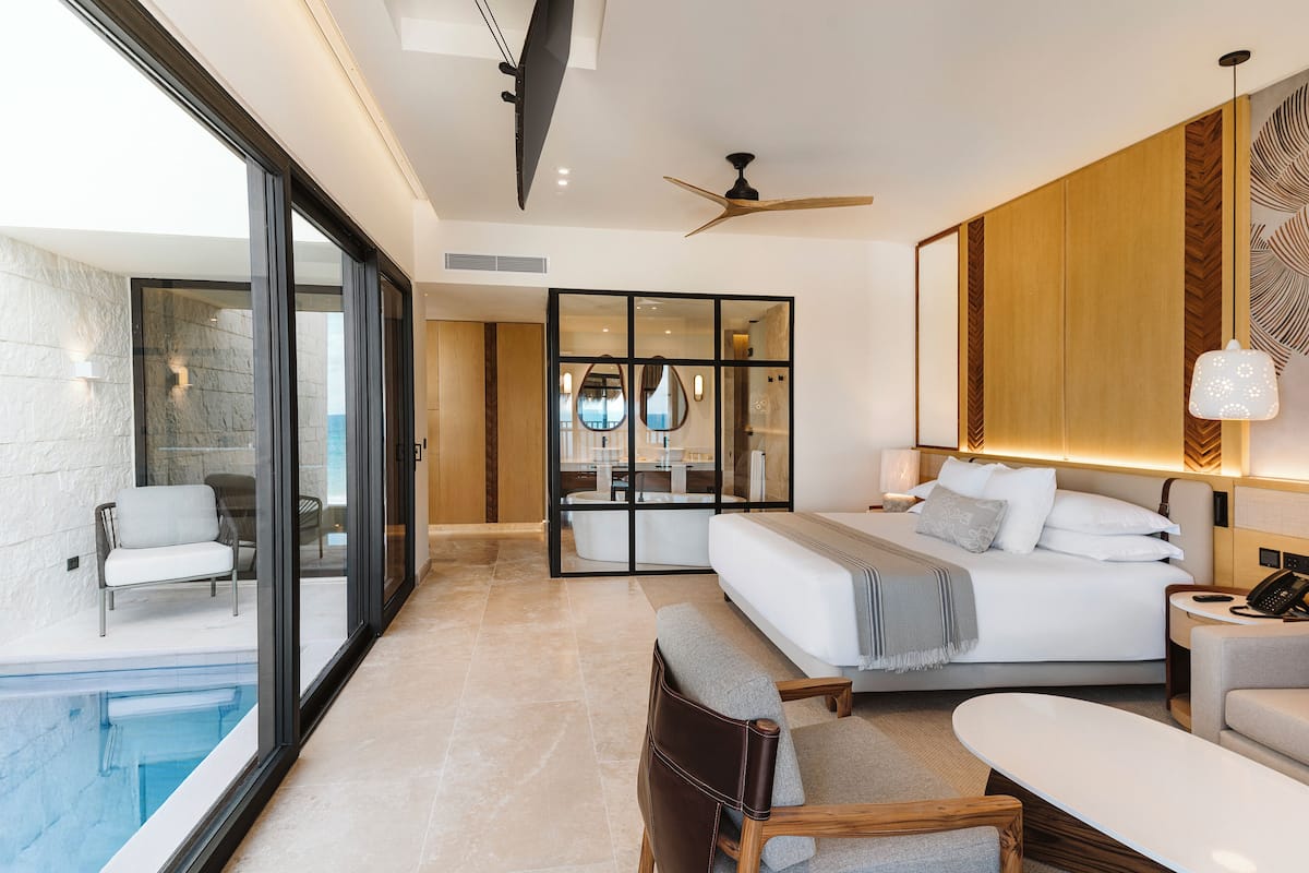 A modern hotel room with a large bed, a seating area, a small pool, and a view of the outdoors.