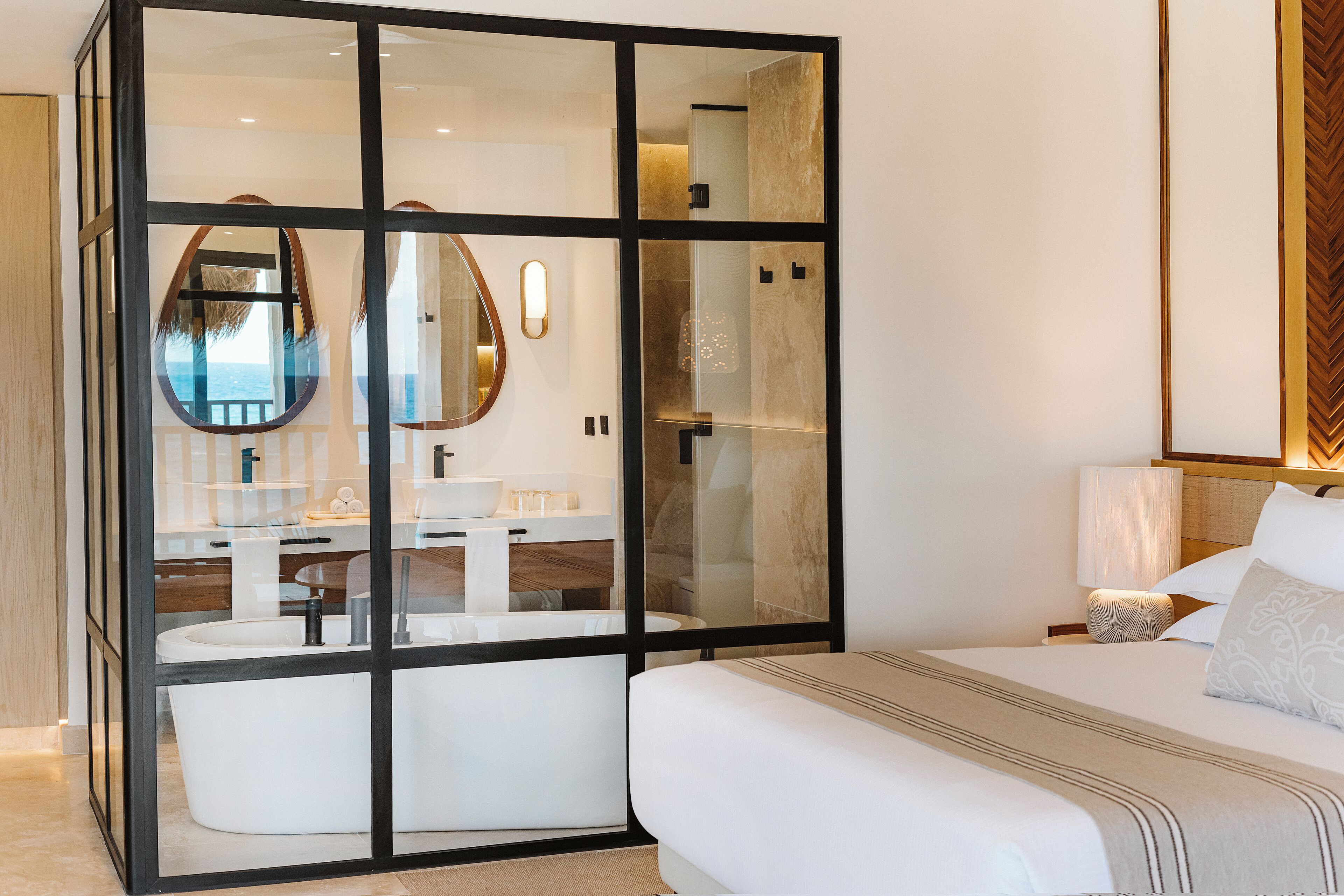 A modern hotel room with a large glass partition, a bed, a bedside table, a lamp, and a view of the sea through the glass.