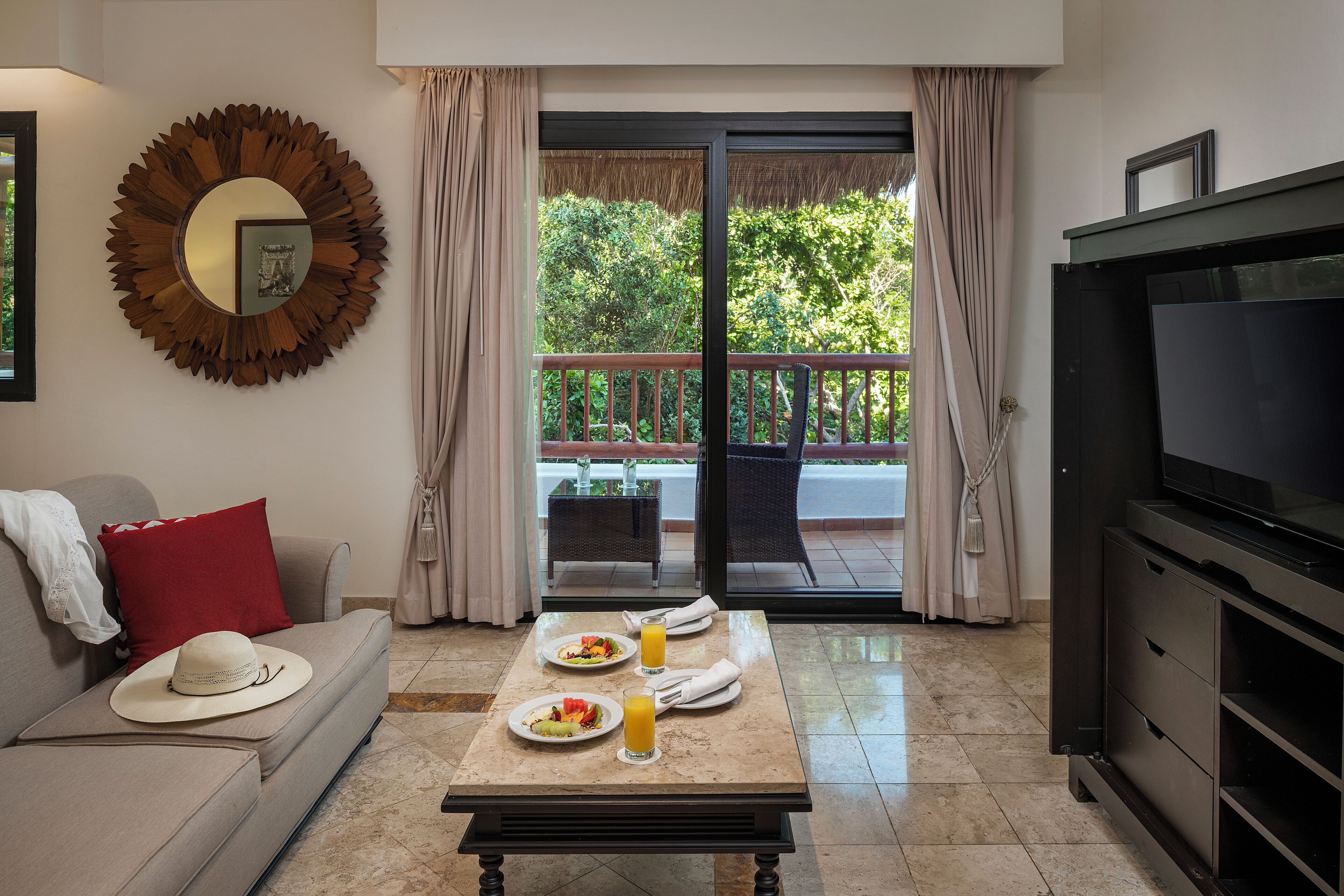A room with a sofa, a dining table with breakfast, a TV, and a balcony with a view of greenery.