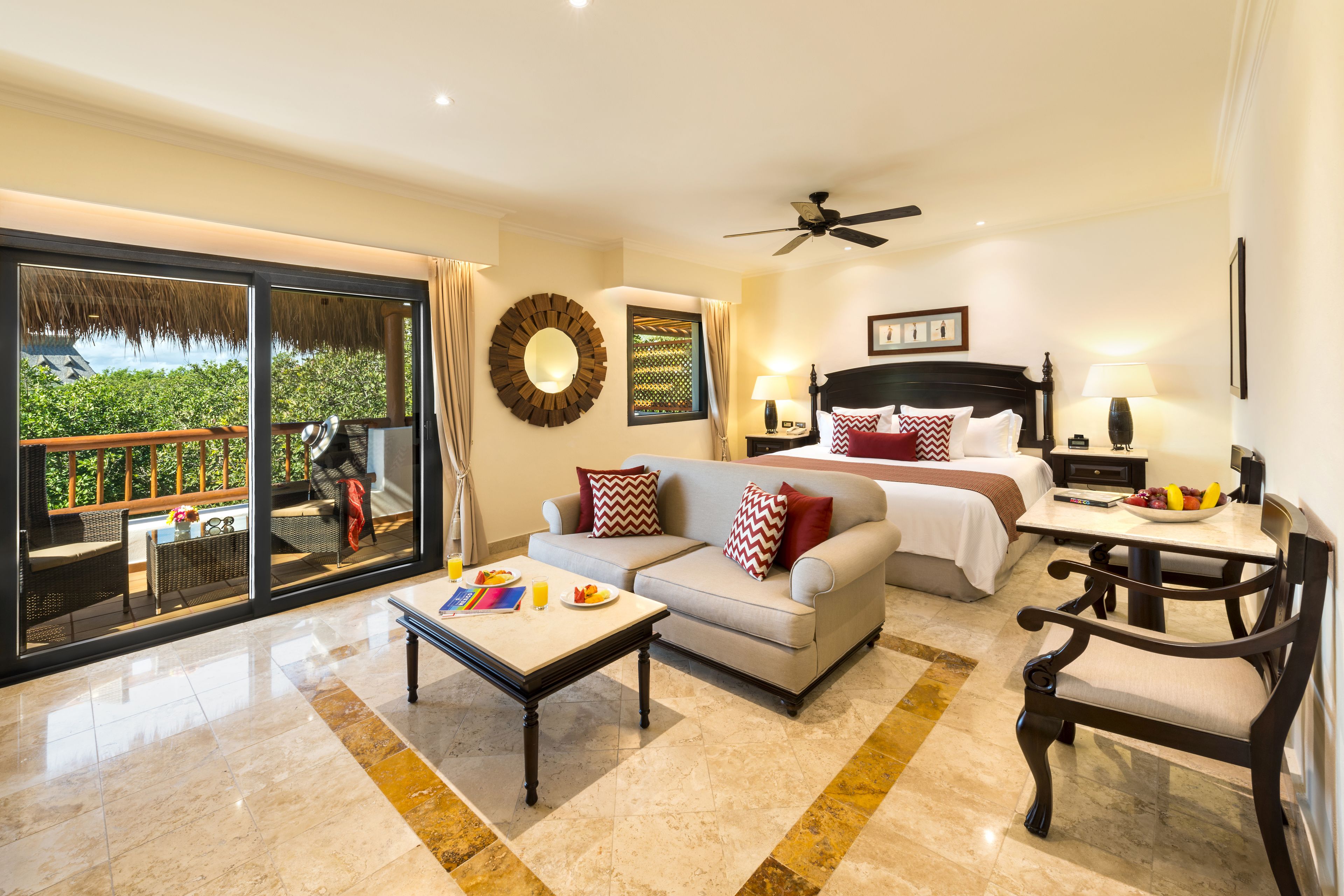 A hotel room with a bed, a sofa, a dining table, and a balcony with a view of greenery.