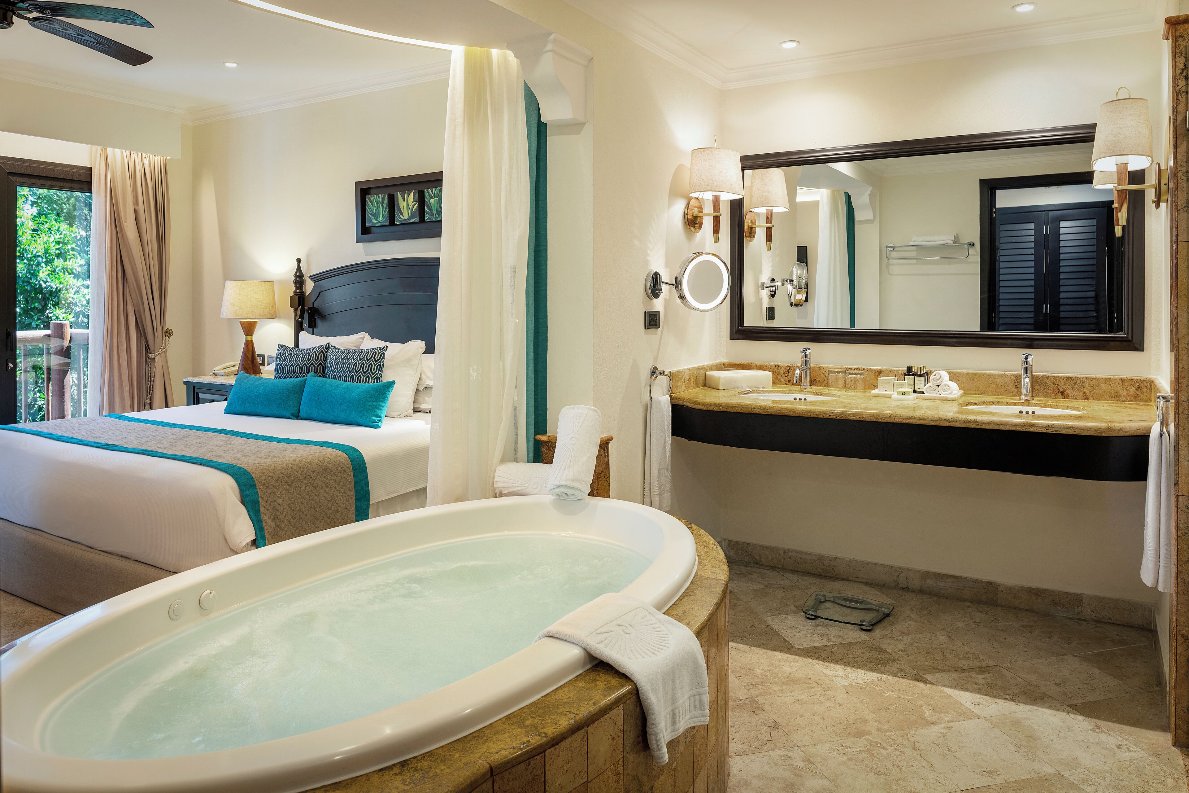 A hotel room with a large bathtub, a bed with teal bedding, a bathroom with a large mirror and sink.
