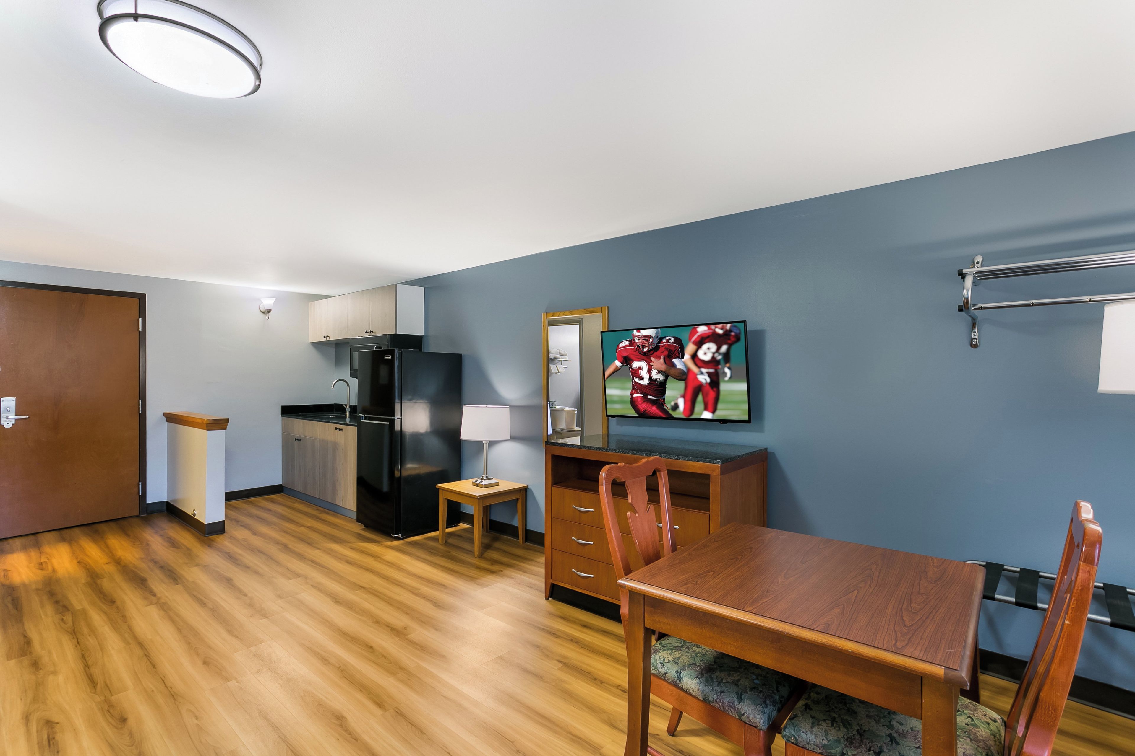 A compact hotel room with a kitchenette, a dining area, and a television displaying a sports event.