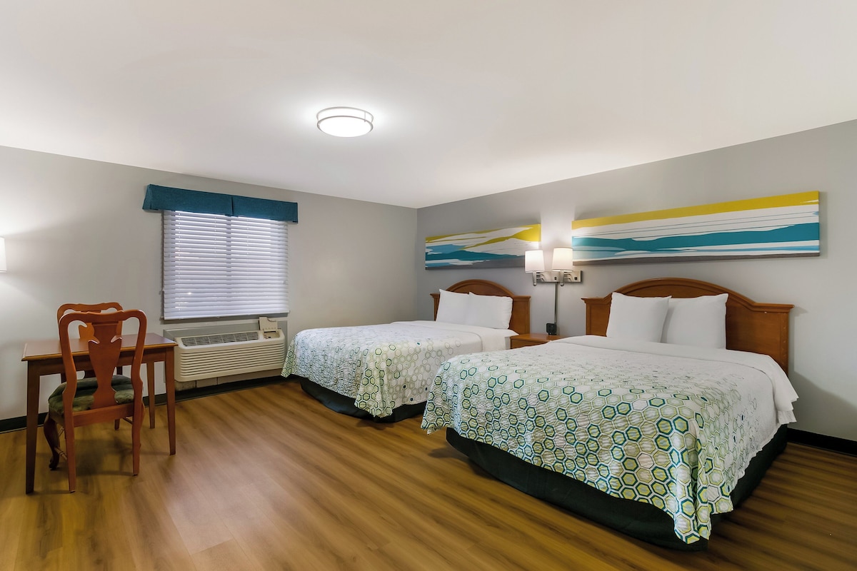 A hotel room with two beds, a wooden chair, a table, a window with blinds, a ceiling light, and a wall painting.
