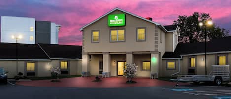 A two-story building with a green sign indicating it's a hotel. There are multiple windows on the facade, and the building is surrounded by a parking area.