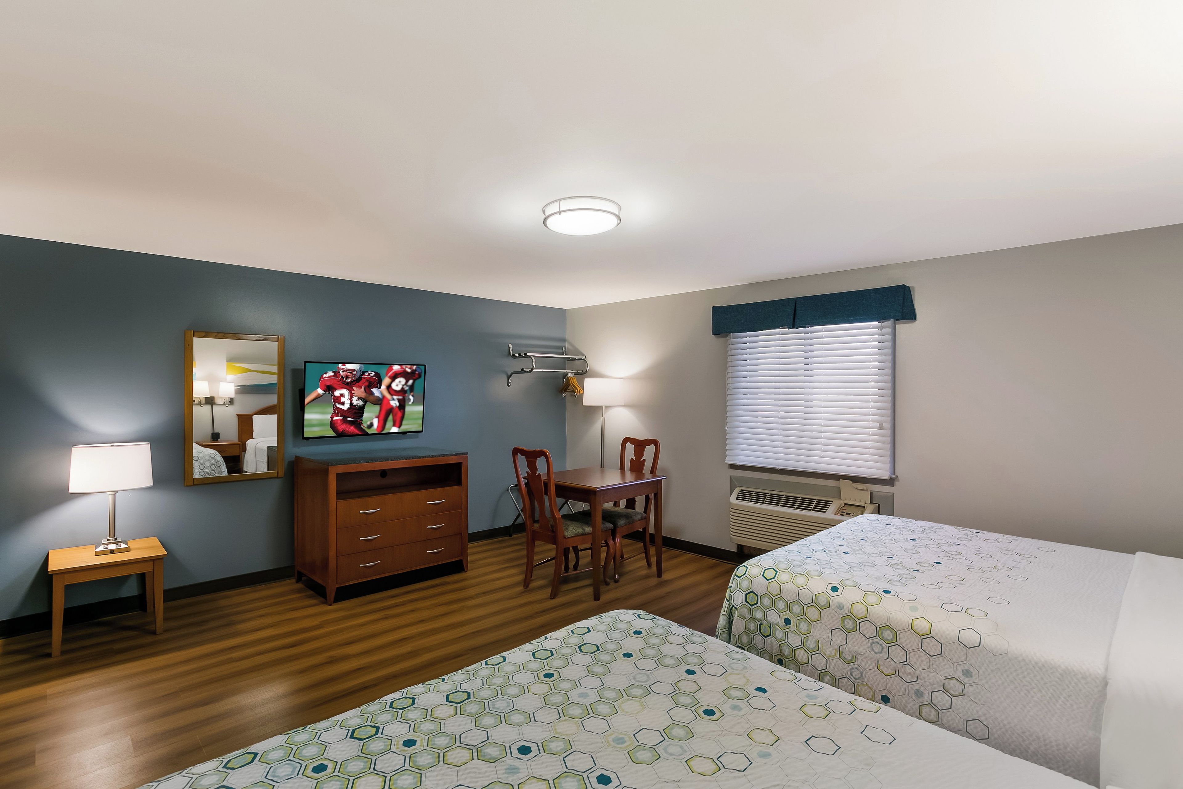 A hotel room with a wooden floor, a desk with a chair, a TV on the wall, a bed with a patterned bedspread, and a window with blinds.