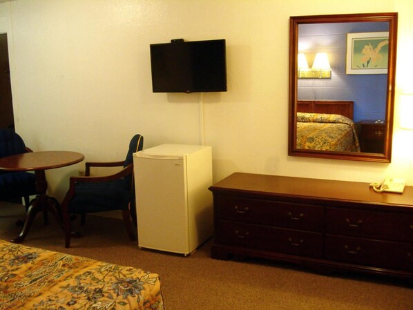 Habitación de hotel con cama, refrigerador pequeño, televisor montado en la pared, cómoda de madera, escritorio con silla y piso alfombrado.