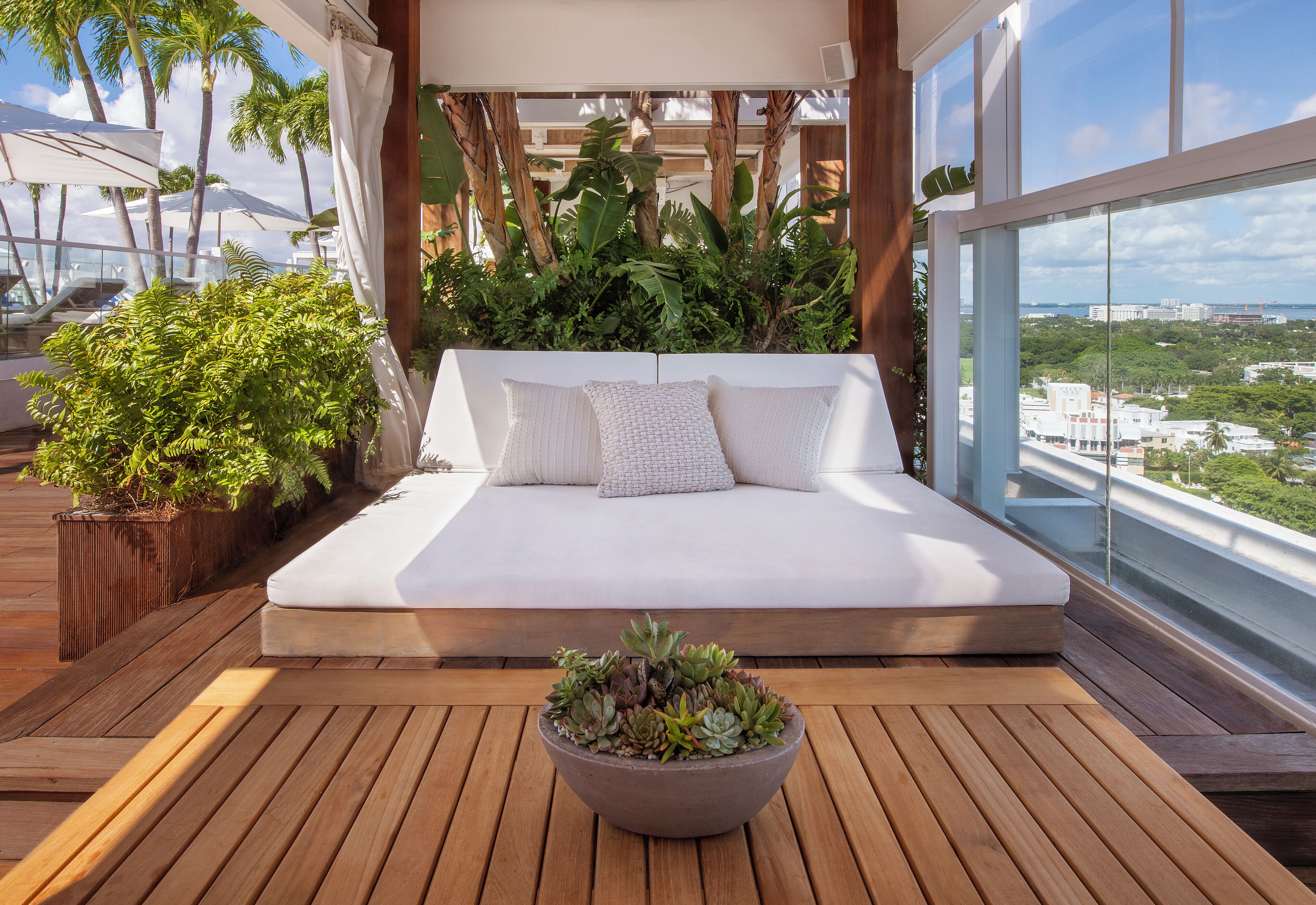 A balcony with a wooden floor, a white sofa with cushions, and a potted plant in the center.