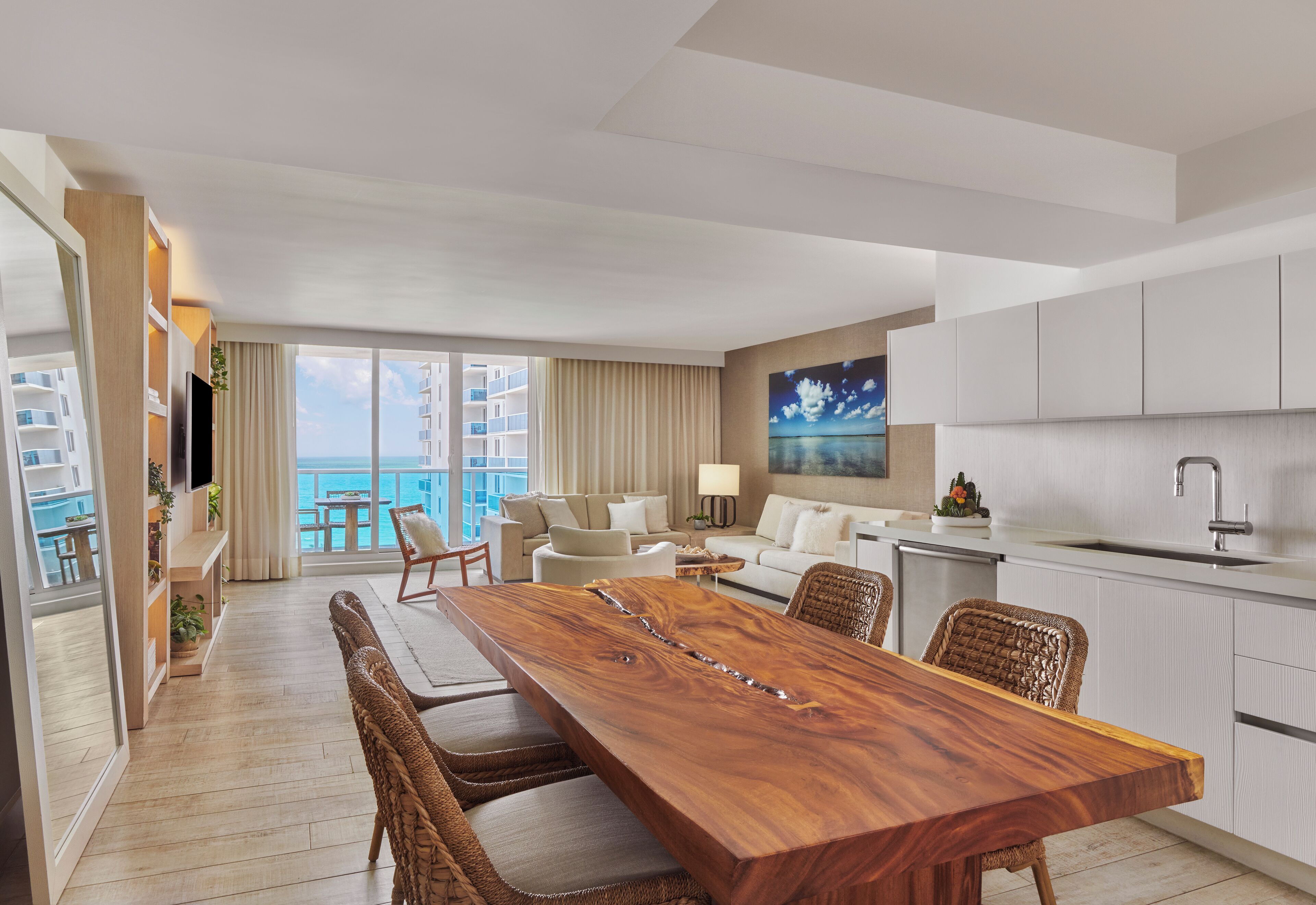 A modern hotel room with a wooden dining table, wicker chairs, a sofa, and a kitchen area.