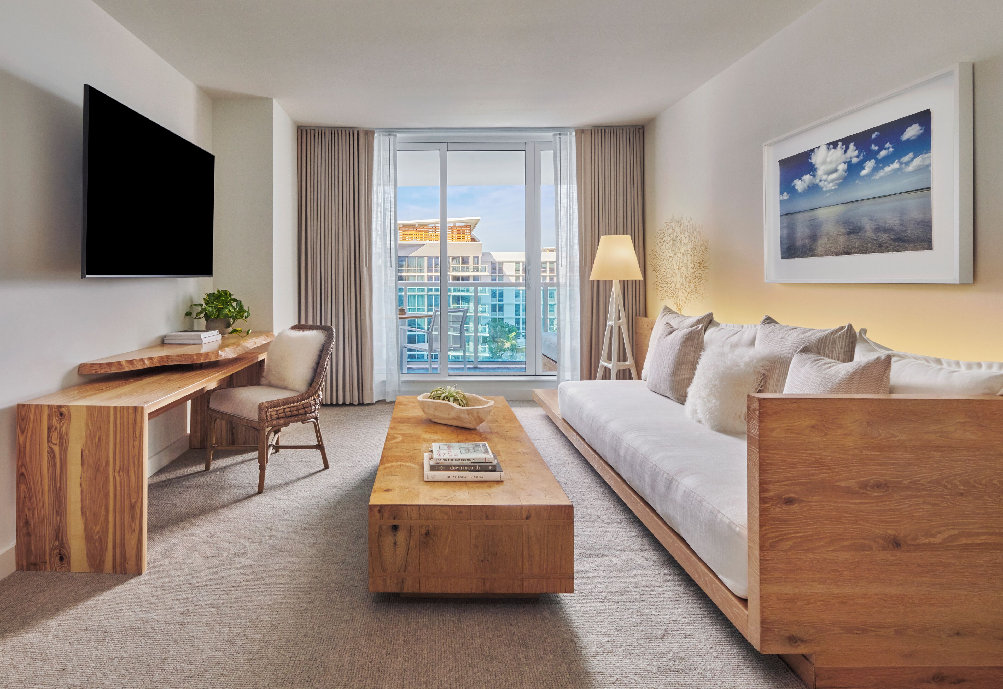 A modern hotel room with a large window, a flat-screen TV, a wooden desk, a comfortable sofa, and a coffee table.
