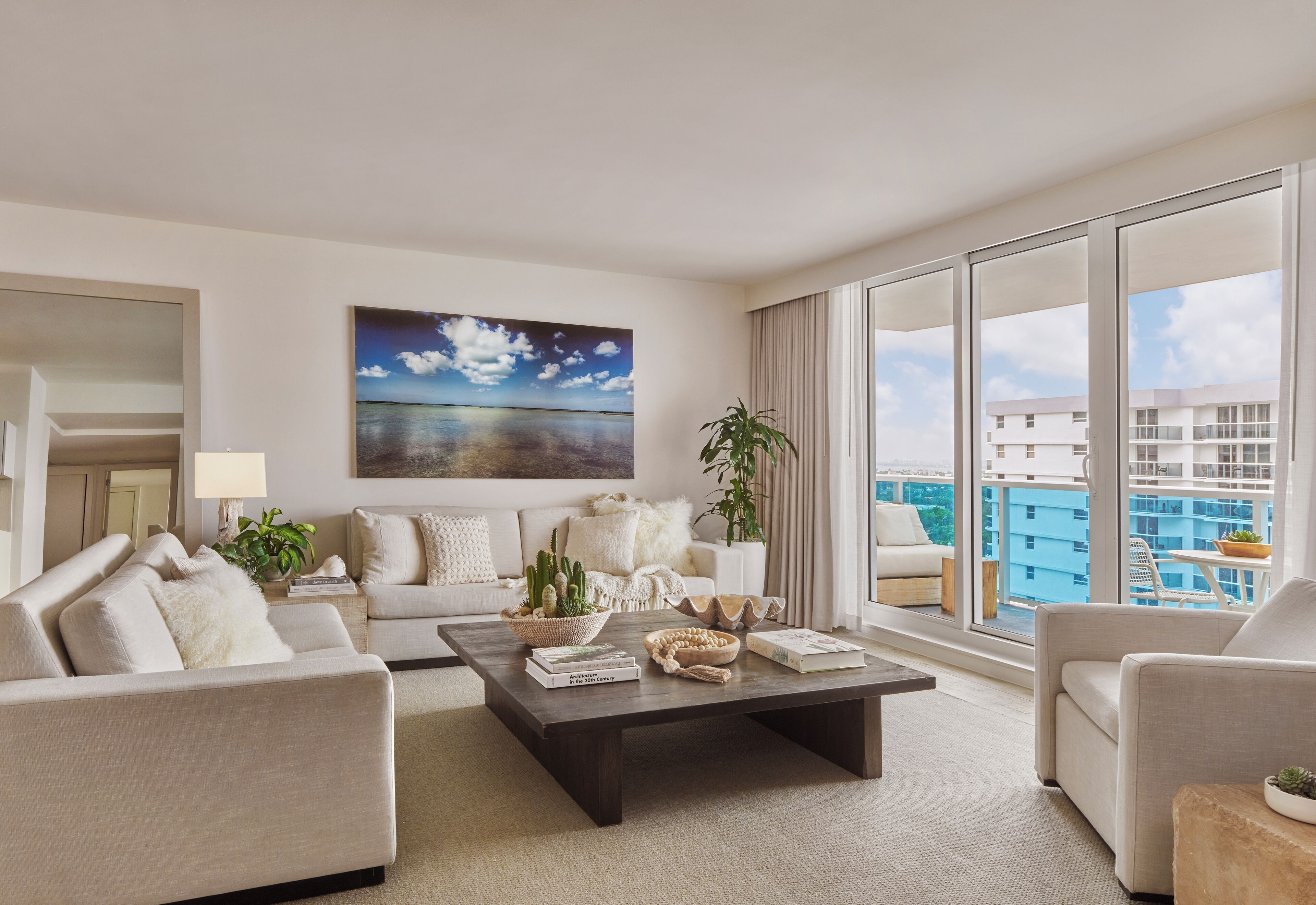 A modern living room with a large window, a painting of a beach, and a view of a cityscape.