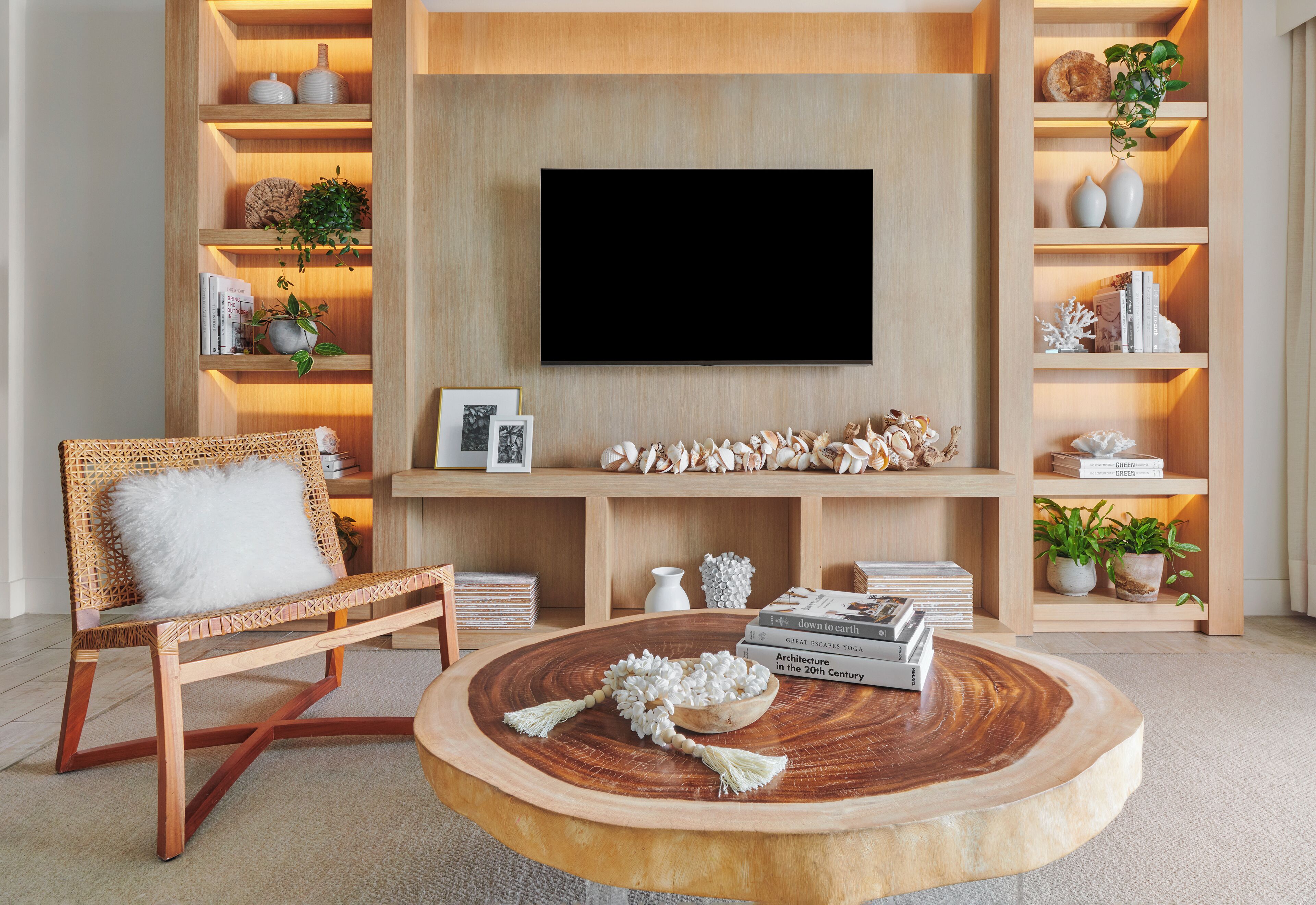 Un salon doté d’un téléviseur à écran plat, d’étagères en bois ornées d’objets décoratifs, d’un fauteuil en osier avec un coussin blanc et d’une table basse ronde en bois sur laquelle sont posés des livres et un vase décoratif.
