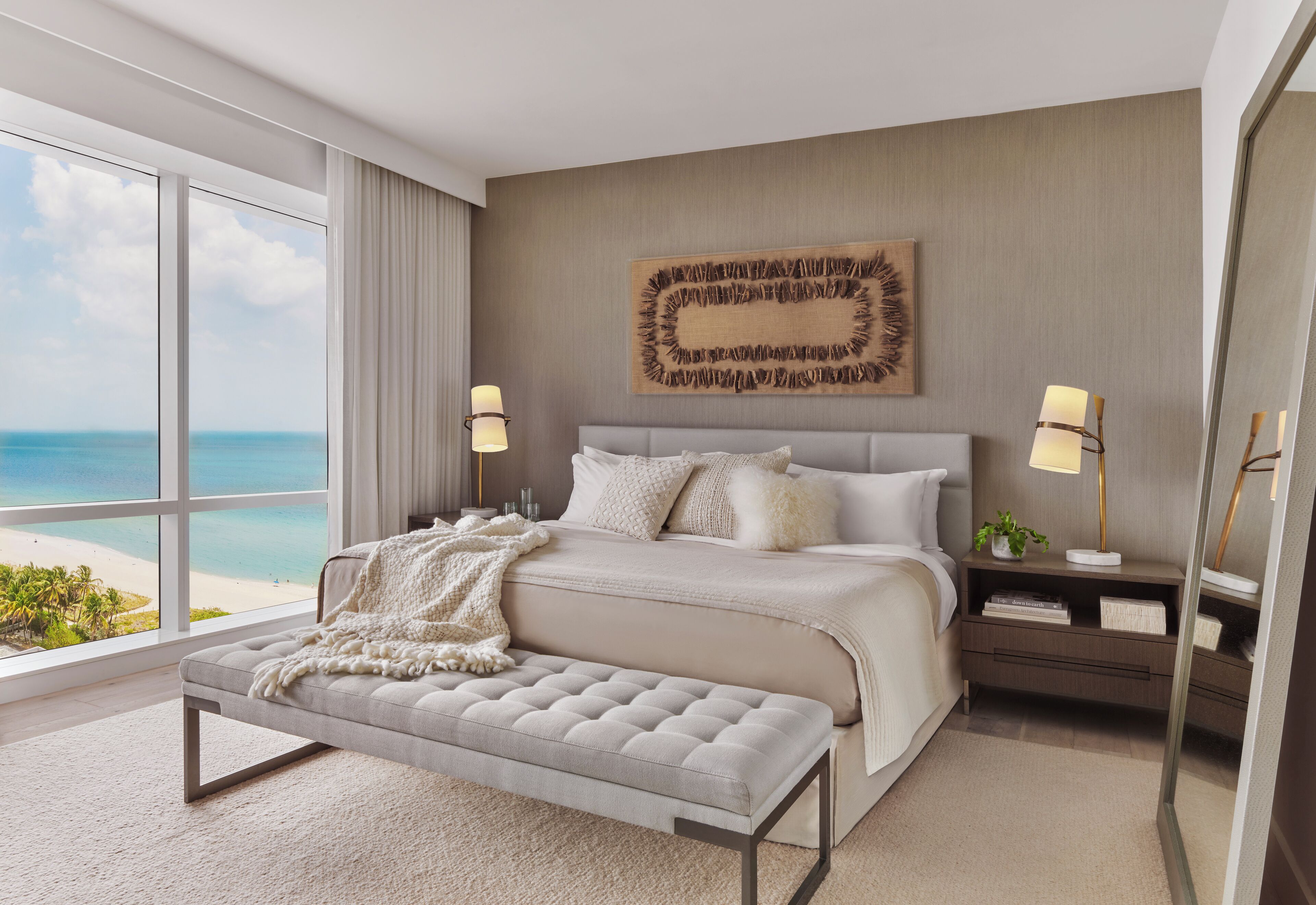 A modern bedroom with a large bed, two bedside tables with lamps, a bench, and a view of the ocean.