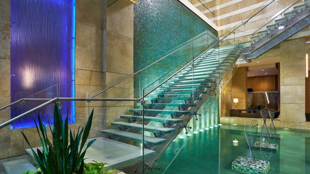 Un escalier intérieur moderne, avec des rampes en verre, une fontaine et un comptoir d’accueil en arrière-plan.