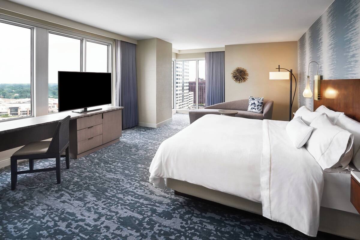 Une chambre d’hôtel dotée d’un grand lit, d’un bureau avec une télévision, d’un canapé et offrant une vue sur la ville grâce à de grandes fenêtres.