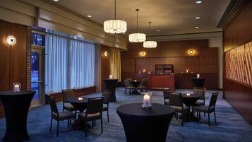 A hotel lobby with round tables, chairs, and hanging pendant lights. There is a reception desk with a counter and a display case.