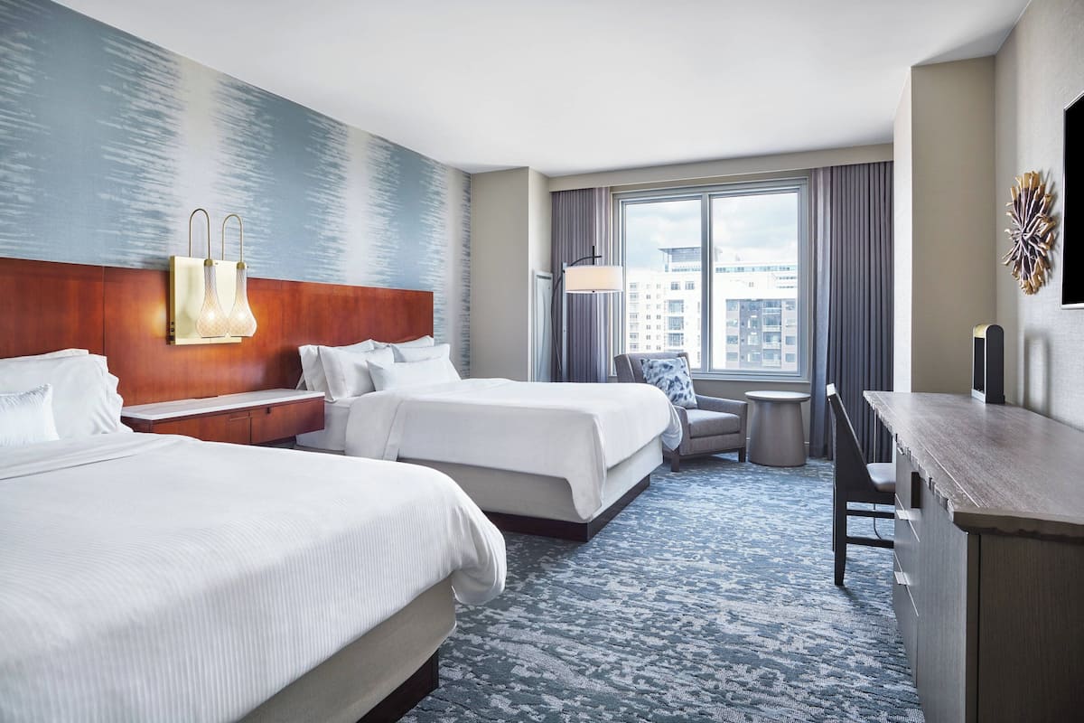 Une chambre d’hôtel avec deux lits, un bureau, une chaise, une lampe et une vue sur la ville grâce à de grandes fenêtres.
