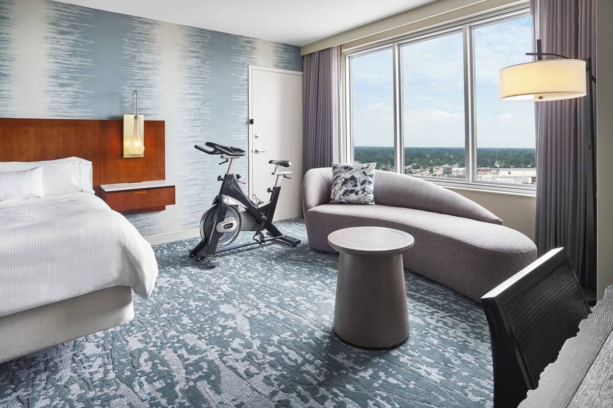 Une chambre d’hôtel avec un lit, un canapé, une petite table ronde, un vélo d’appartement et une grande fenêtre donnant sur un beau paysage.