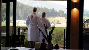 Un couple, vêtu de longues robes blanches, se tient près d’une fenêtre donnant sur un paysage magnifique.