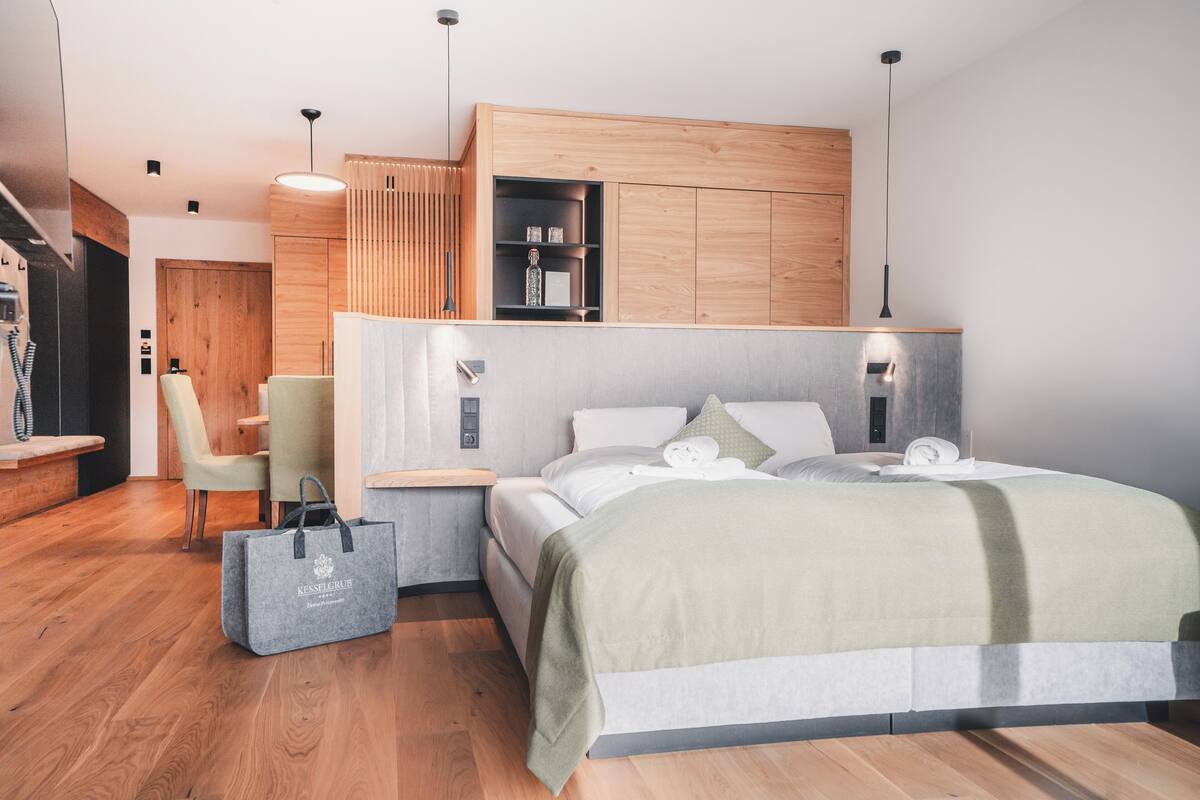 A modern hotel room with a wooden bed frame, a desk with a chair, a wardrobe, and a hanging light fixture.