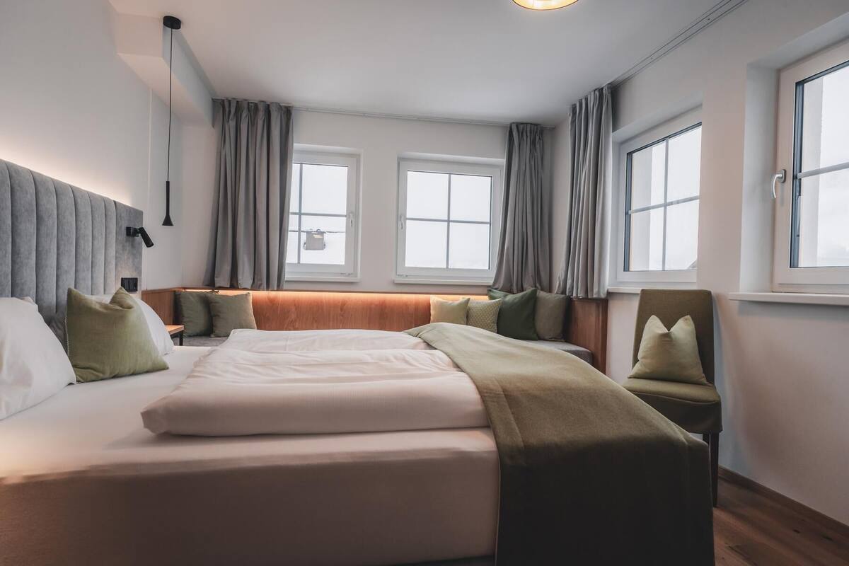 A hotel room with a large bed, two windows, a chair, and a bedside table.