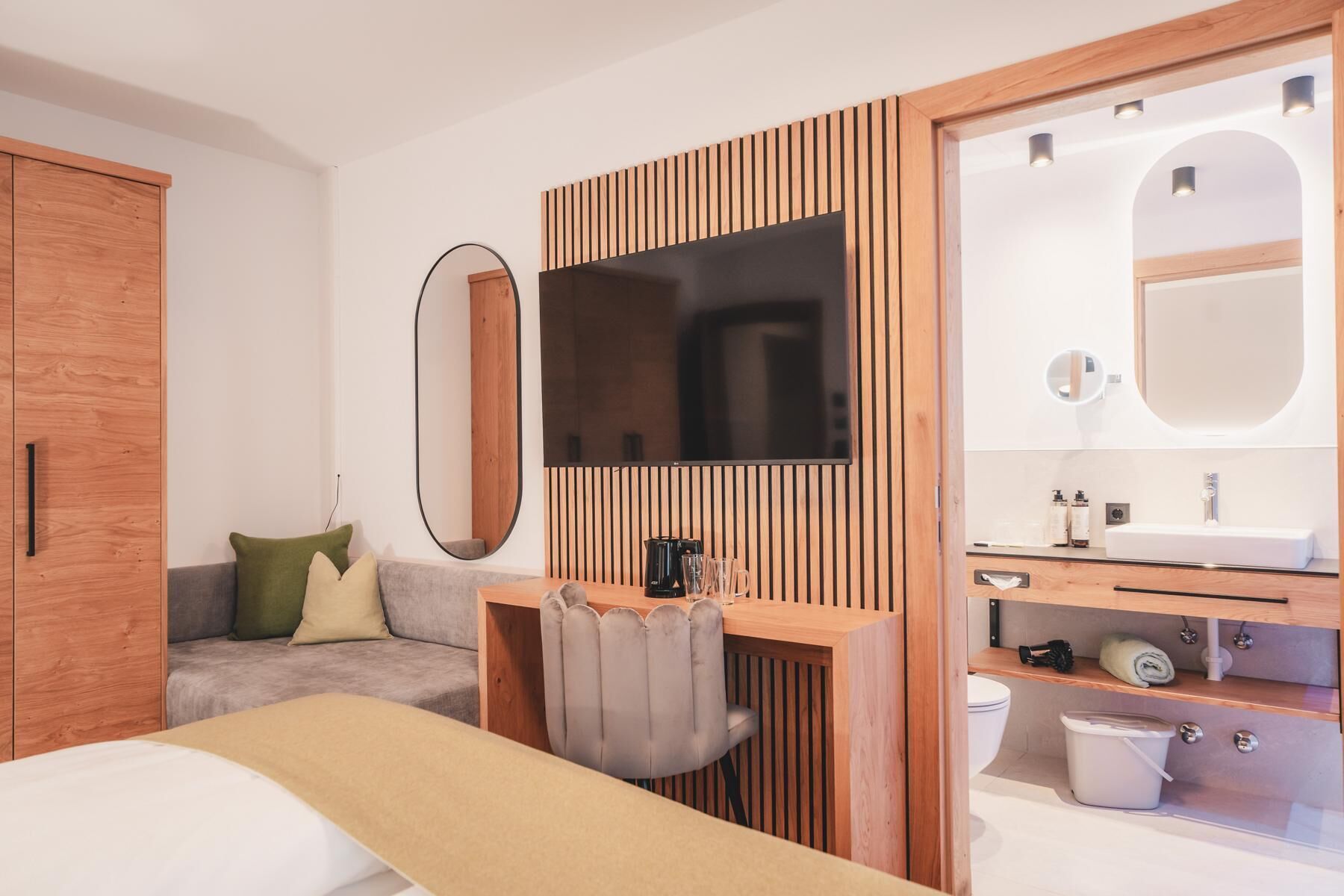 Ein modernes Hotelzimmer mit einem Holzschrank, einem grauen Sofa, einem kleinen Schreibtisch mit Stuhl, einem Flachbildfernseher, einem großen Spiegel und einem Badezimmer mit Waschbecken und Toilette.