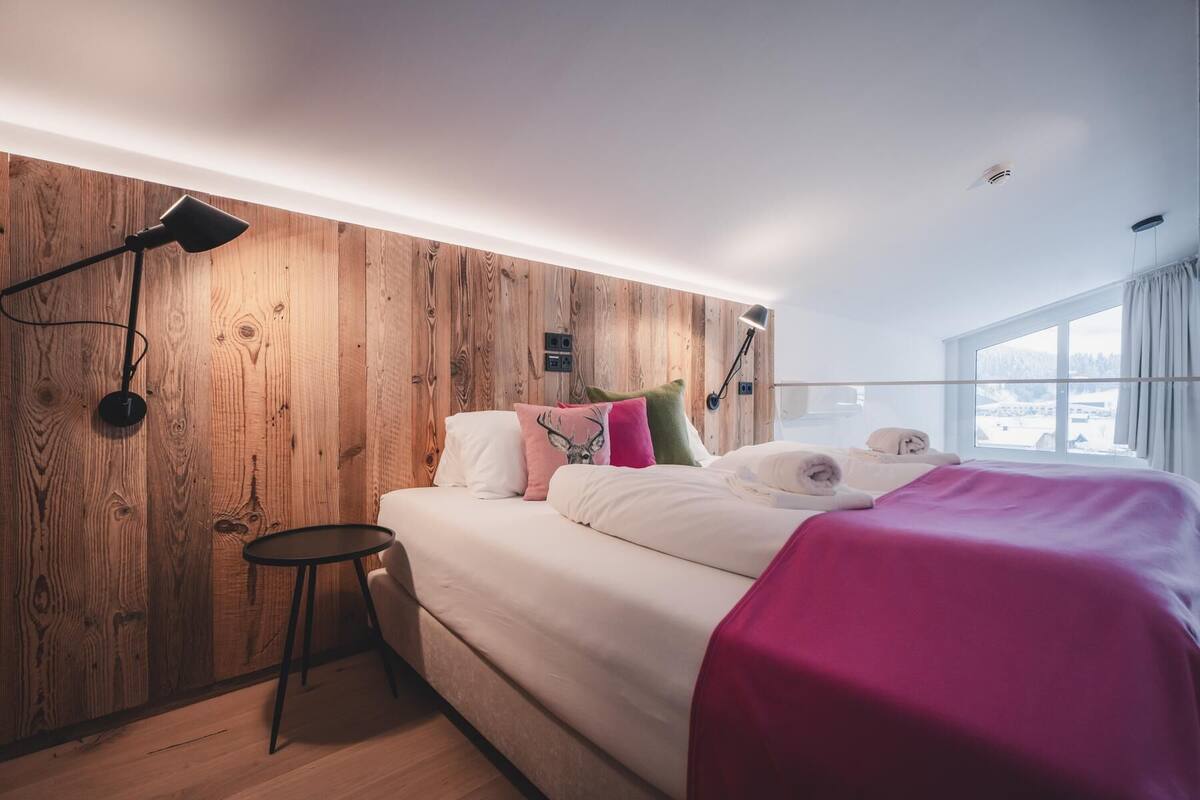 A modern bedroom with a wooden headboard, a bed with white bedding and colorful pillows, a small round table, and a large window with a view of a snowy landscape.