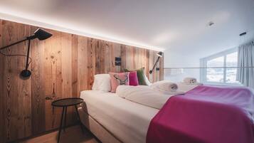 A modern bedroom with a wooden headboard, a bed with white bedding and colorful pillows, a small round table, and a large window with a view of a snowy landscape.