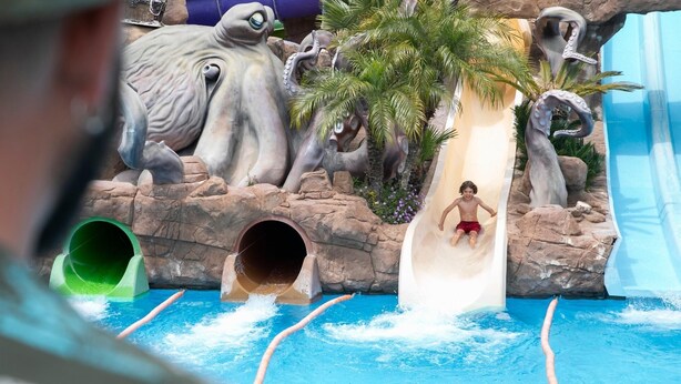 A water slide with a child at the bottom, surrounded by rock formations and palm trees. - Evenia Olympic Palace (Lloret de Mar)