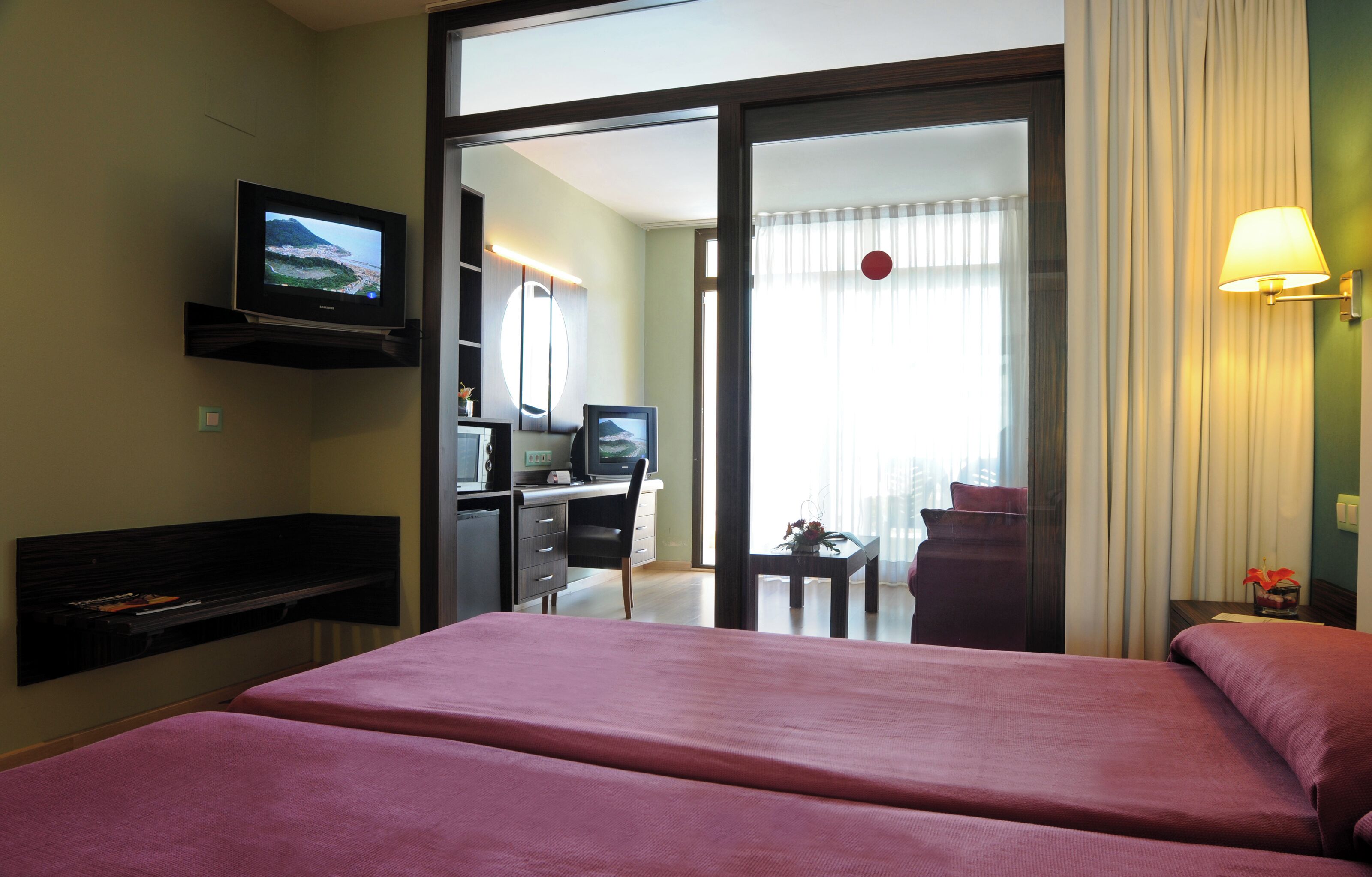 Habitación de hotel con cama, escritorio con computadora, televisor y puerta corrediza a un balcón.