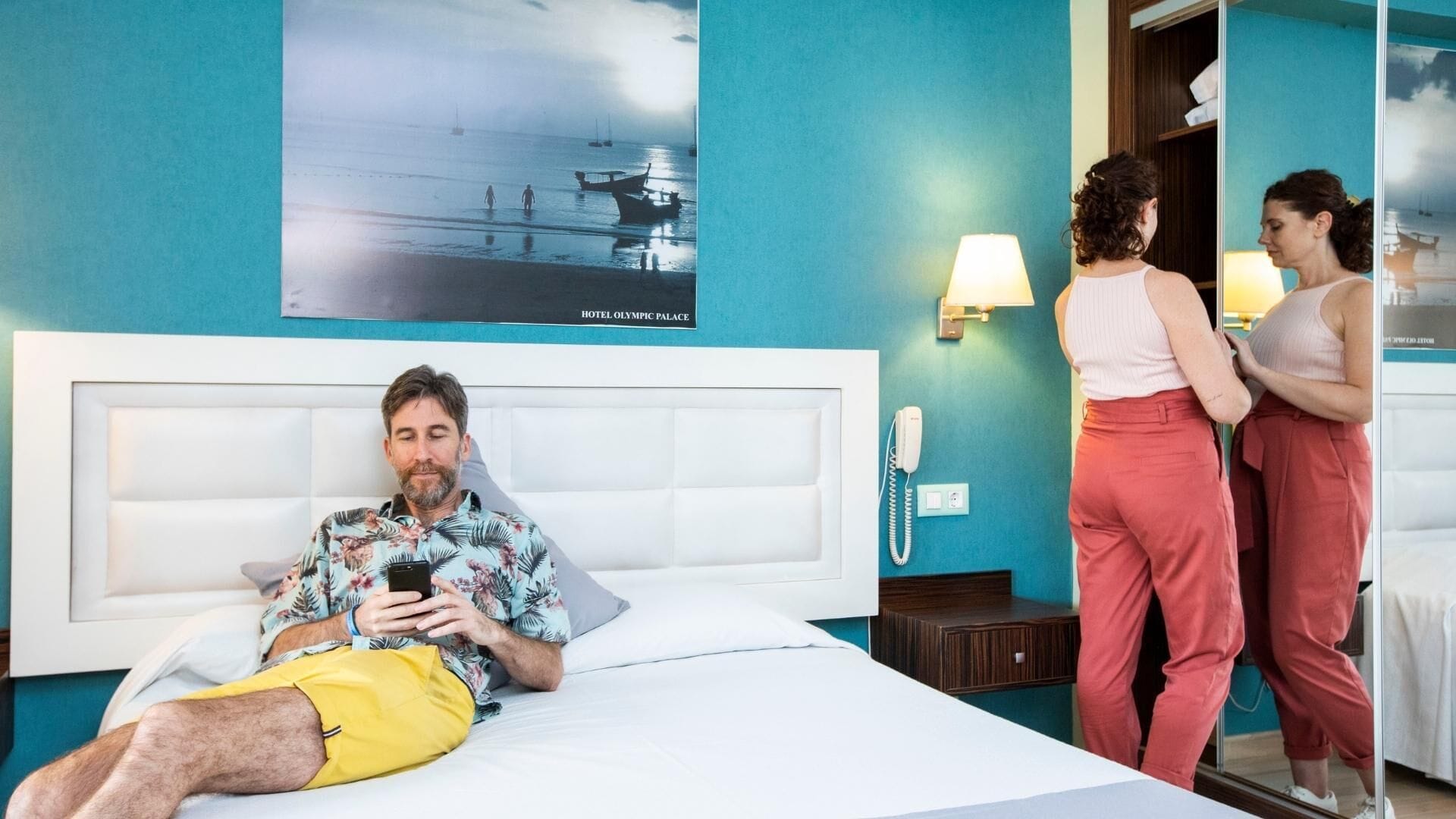 A hotel room with a bed, a bedside phone, and a poster of a beach scene.