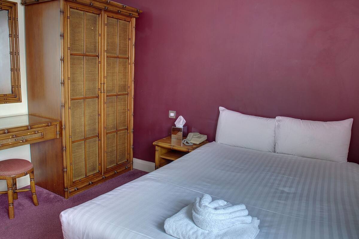 A hotel room with a bed, a wooden wardrobe, a desk with a chair, and a nightstand with a phone.