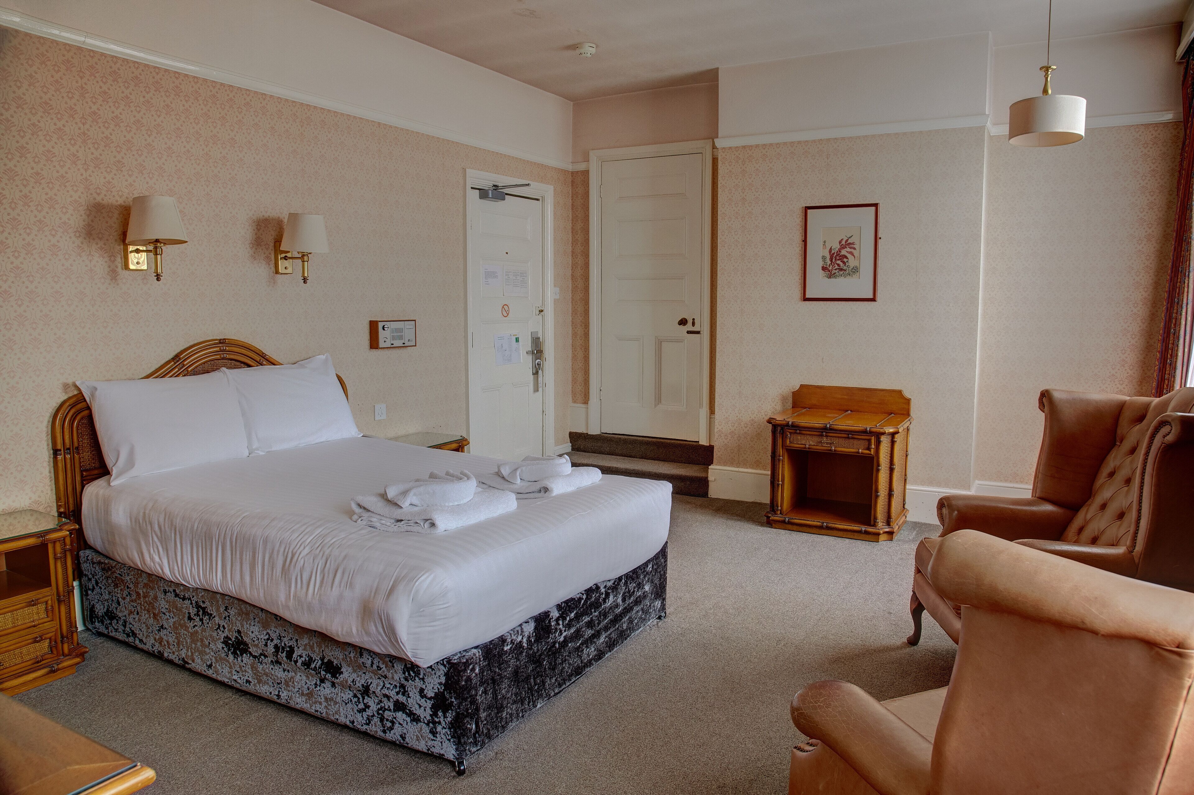 A neatly made bed with white linens and towels, a wooden nightstand, a brown leather armchair, and a small wooden table with a lamp.