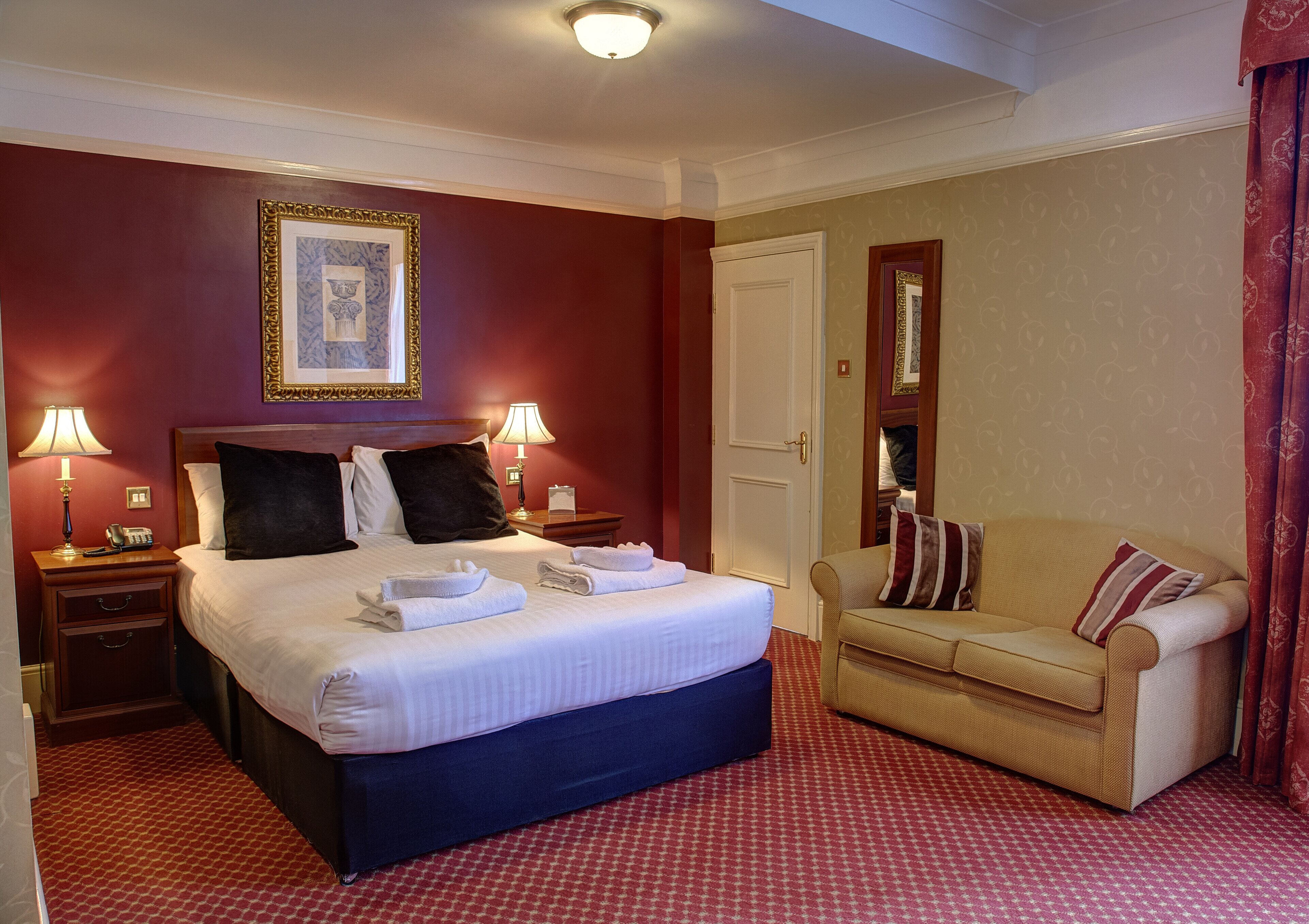 A hotel room with a large bed, two bedside tables with lamps, a sofa, and a door.