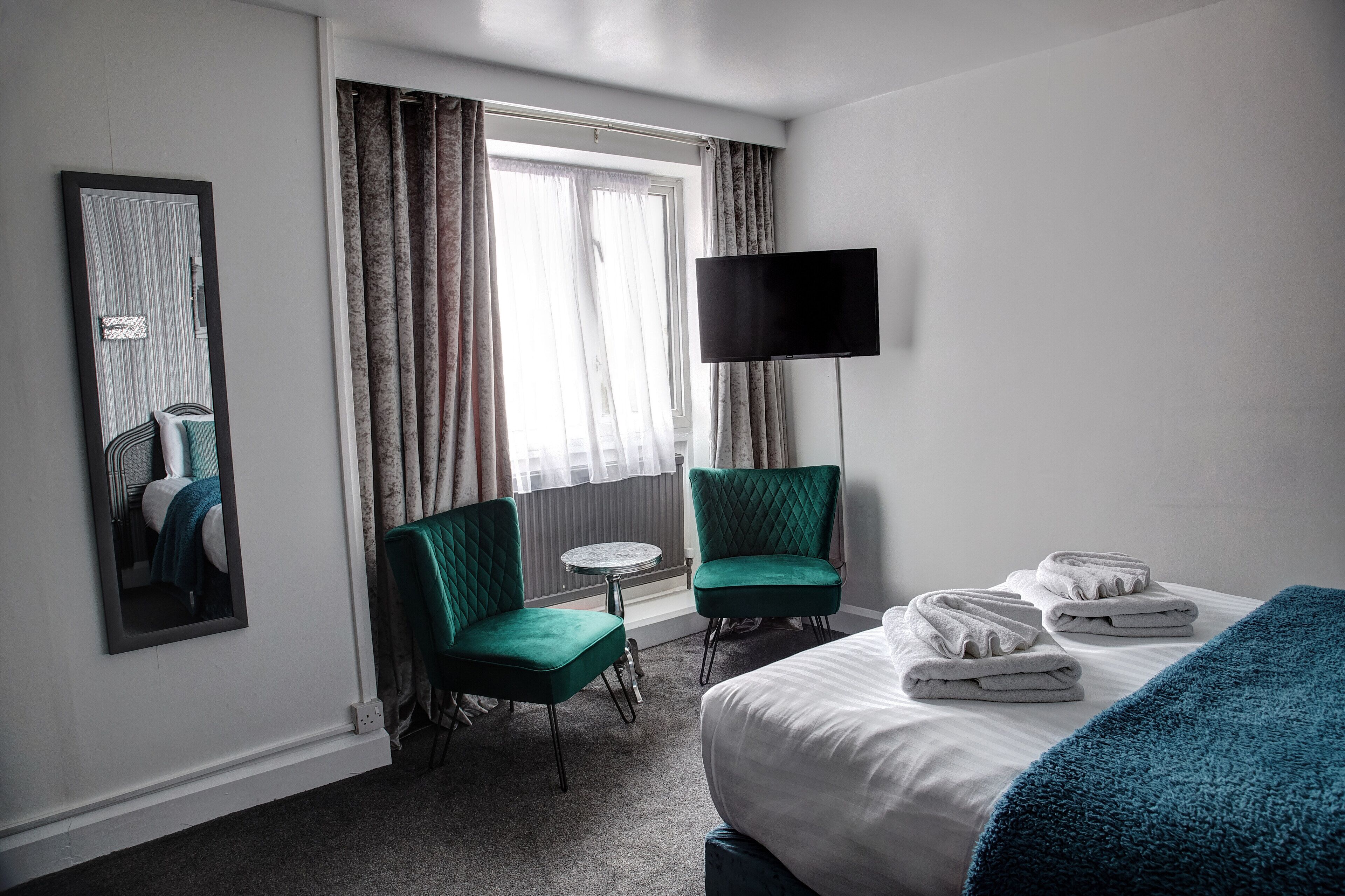 Une chambre d’hôtel avec un lit, deux fauteuils verts, une petite table ronde, un téléviseur à écran plat et une fenêtre avec des rideaux.