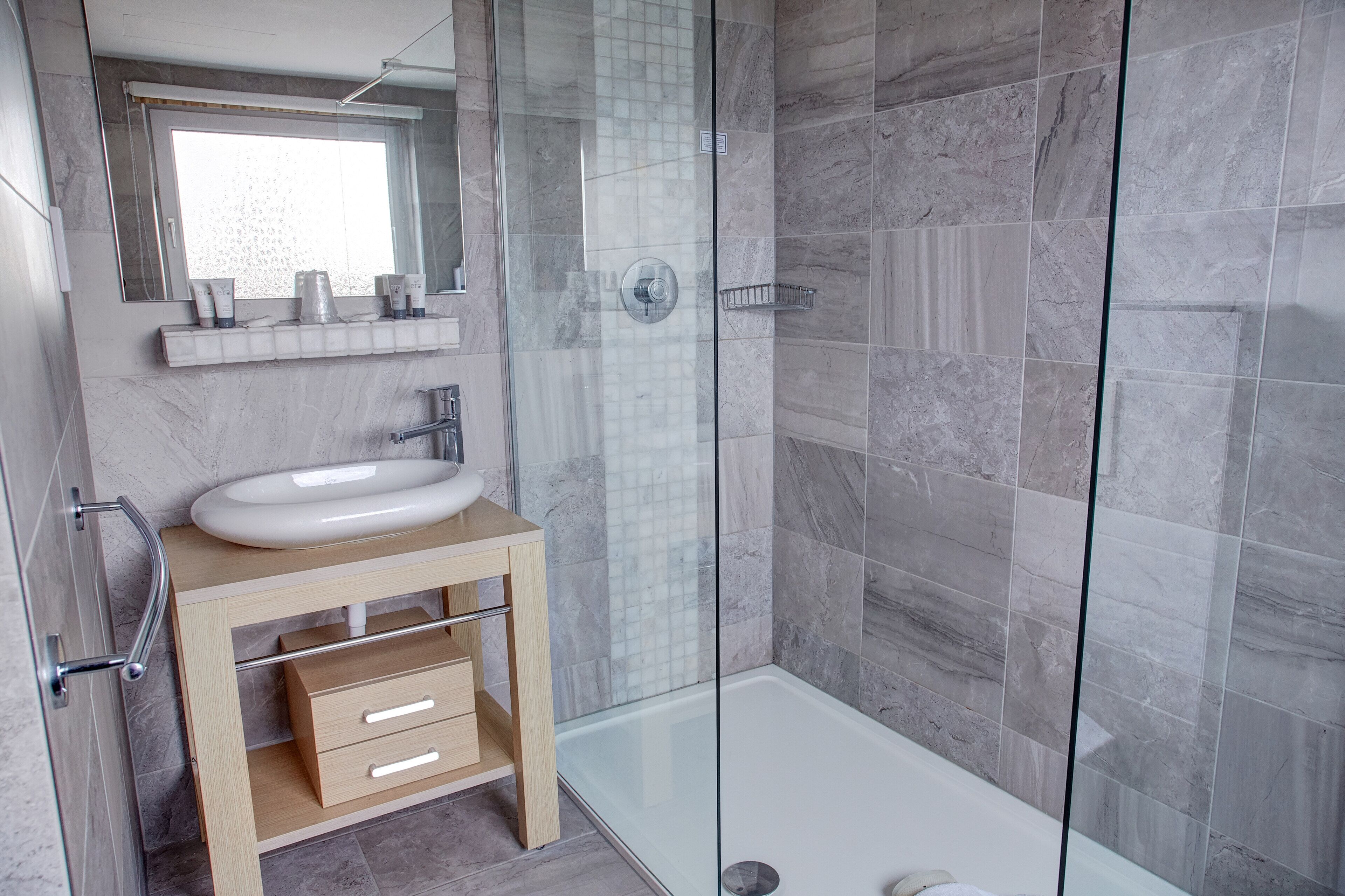 A modern bathroom with a glass-enclosed shower, a marble-tiled wall, a wooden vanity with a sink, and a mirror above it.