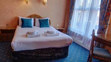 A neatly made bed with white linens and teal pillows, a wooden nightstand with a lamp, and a chair with a floral cushion.